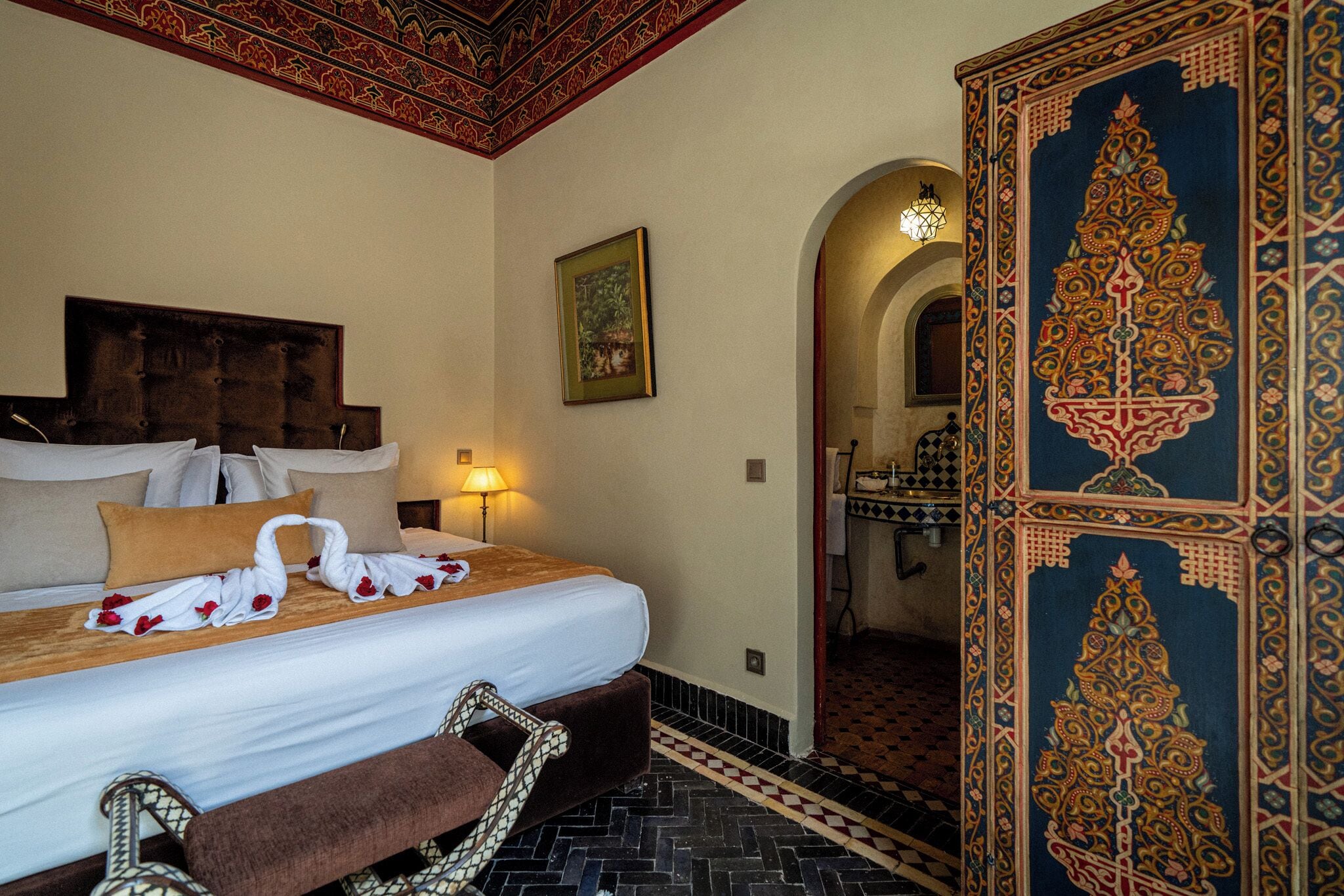 Habitación de hotel con cama, un banco, un espejo y un baño visible a través de un arco.