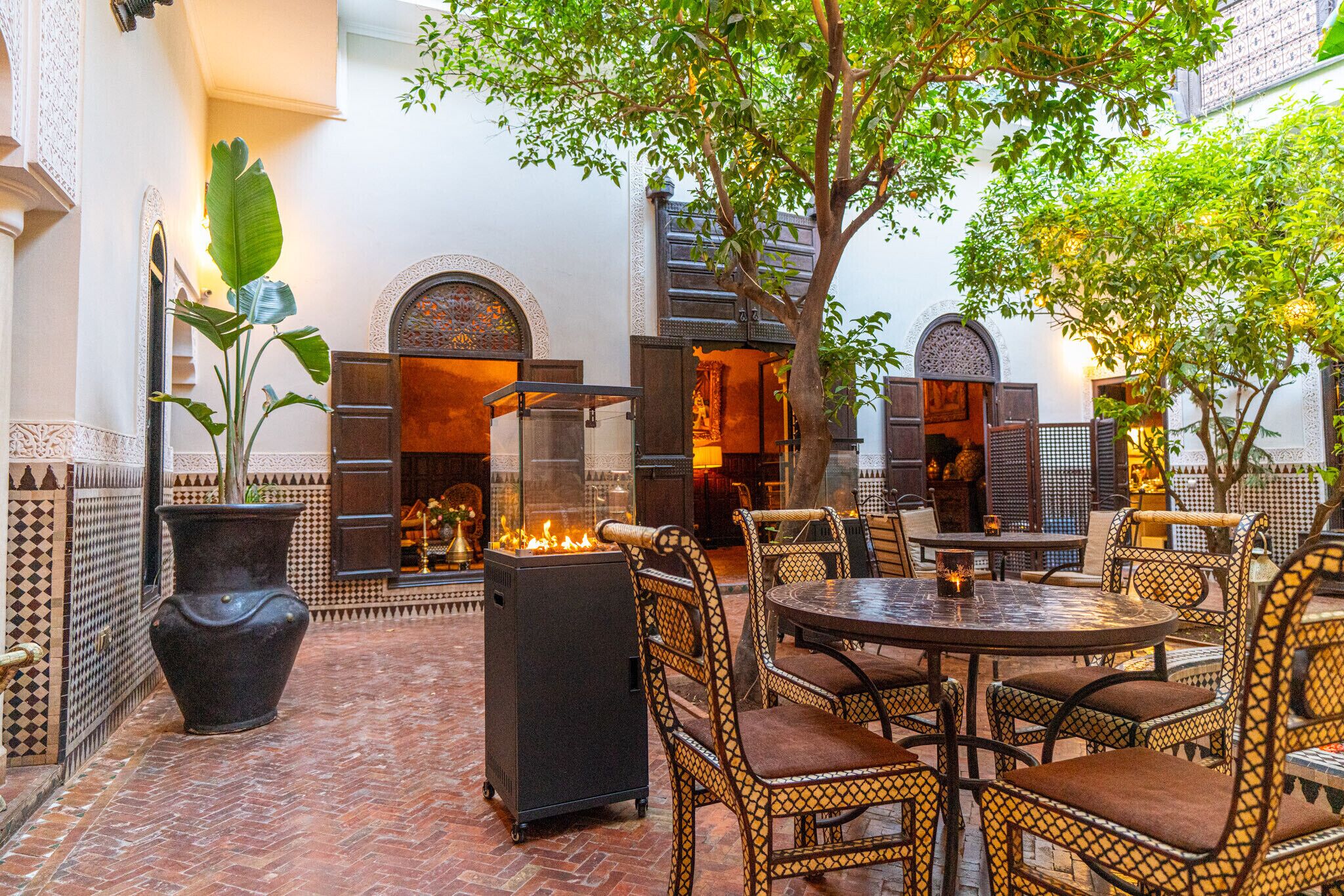 Un patio al aire libre con una mesa redonda y sillas de mimbre, rodeado de plantas en macetas y una chimenea.