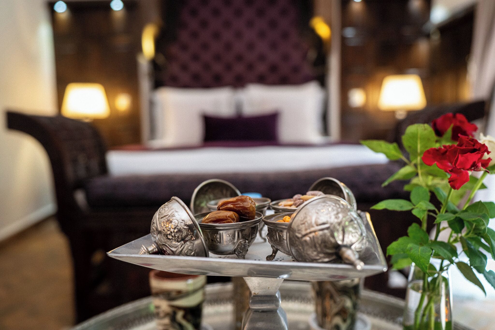 A hotel room with a bed, bedside lamps, and a tray with silverware and a vase with a red flower.