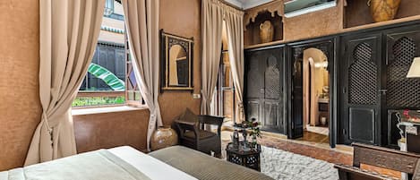 A room with a bed, a chair, a small table with decorative items, and a view of an outdoor area with a gate and greenery.