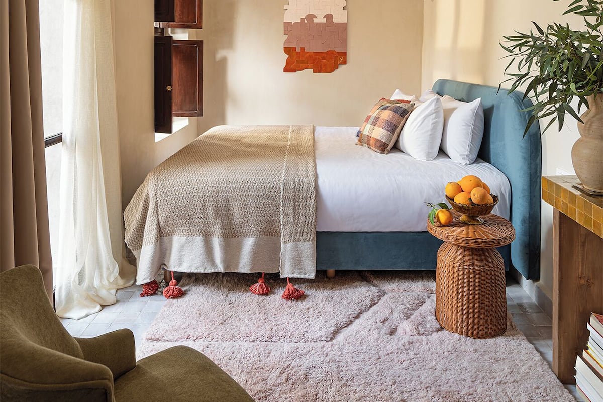 A bedroom with a bed, a blue sofa, a wicker side table with fruit, a potted plant, and a decorative wall piece.