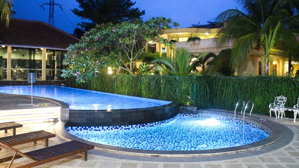 Une piscine dont le bord est recouvert de carreaux, entourée de palmiers et bordée d’un bâtiment dont le toit est également recouvert de carreaux.