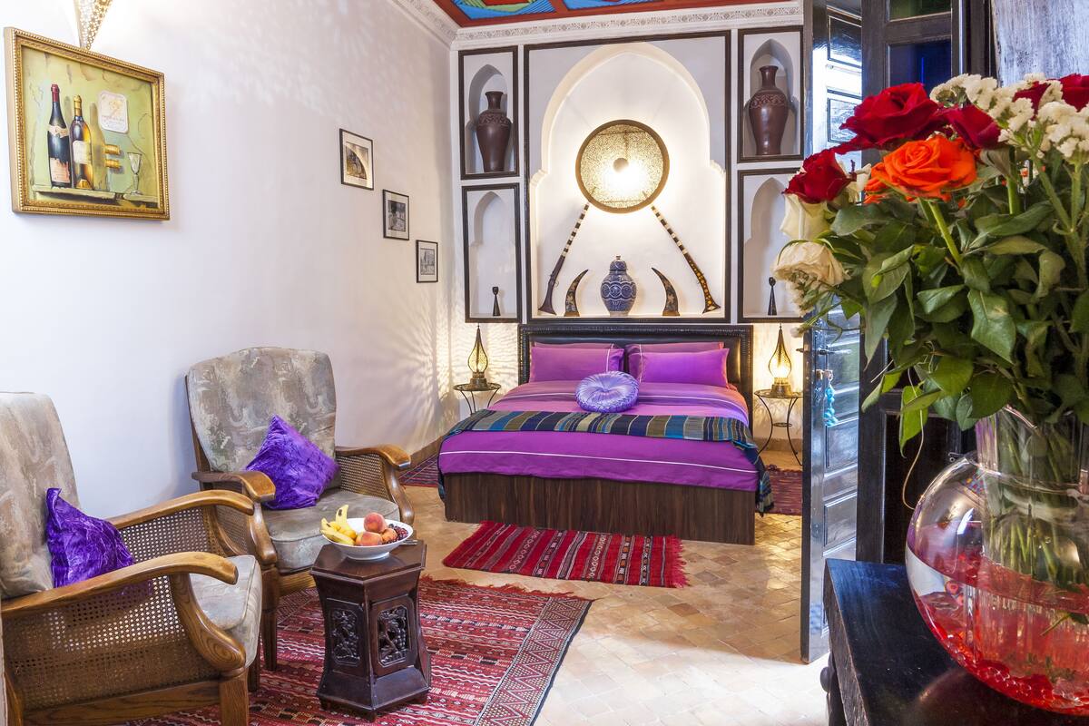 Une pièce comprenant un lit, une chaise, une petite table avec des fruits et un vase de fleurs.