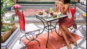 Una mujer sentada en una mesa con una bebida y comida, en un patio con suelo de baldosas y plantas en macetas.