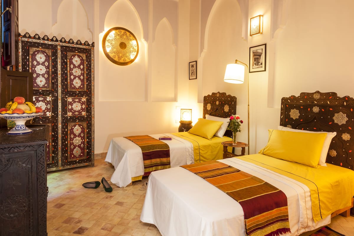 Une chambre d’hôtel avec deux lits, un mur décoratif et un panier de fruits posé sur une commode.