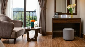 A modern room with a large window, a sofa, a small table with a vase of flowers, a mirror, and a wooden cabinet. - Tam Coc Nam Hoa Hotel and Spa (Nam Hoa Lu)