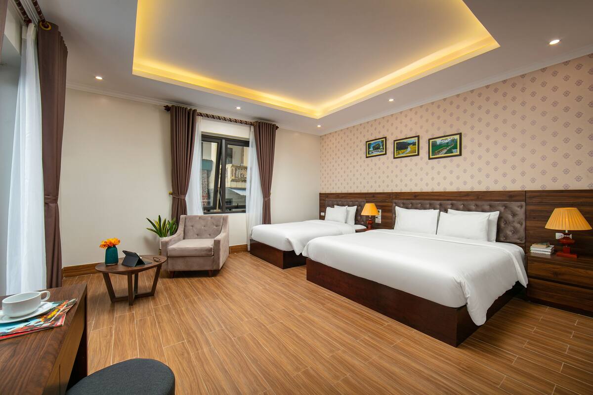 A hotel room with two beds, a sofa, a small table with a cup and a book, and a window with curtains.