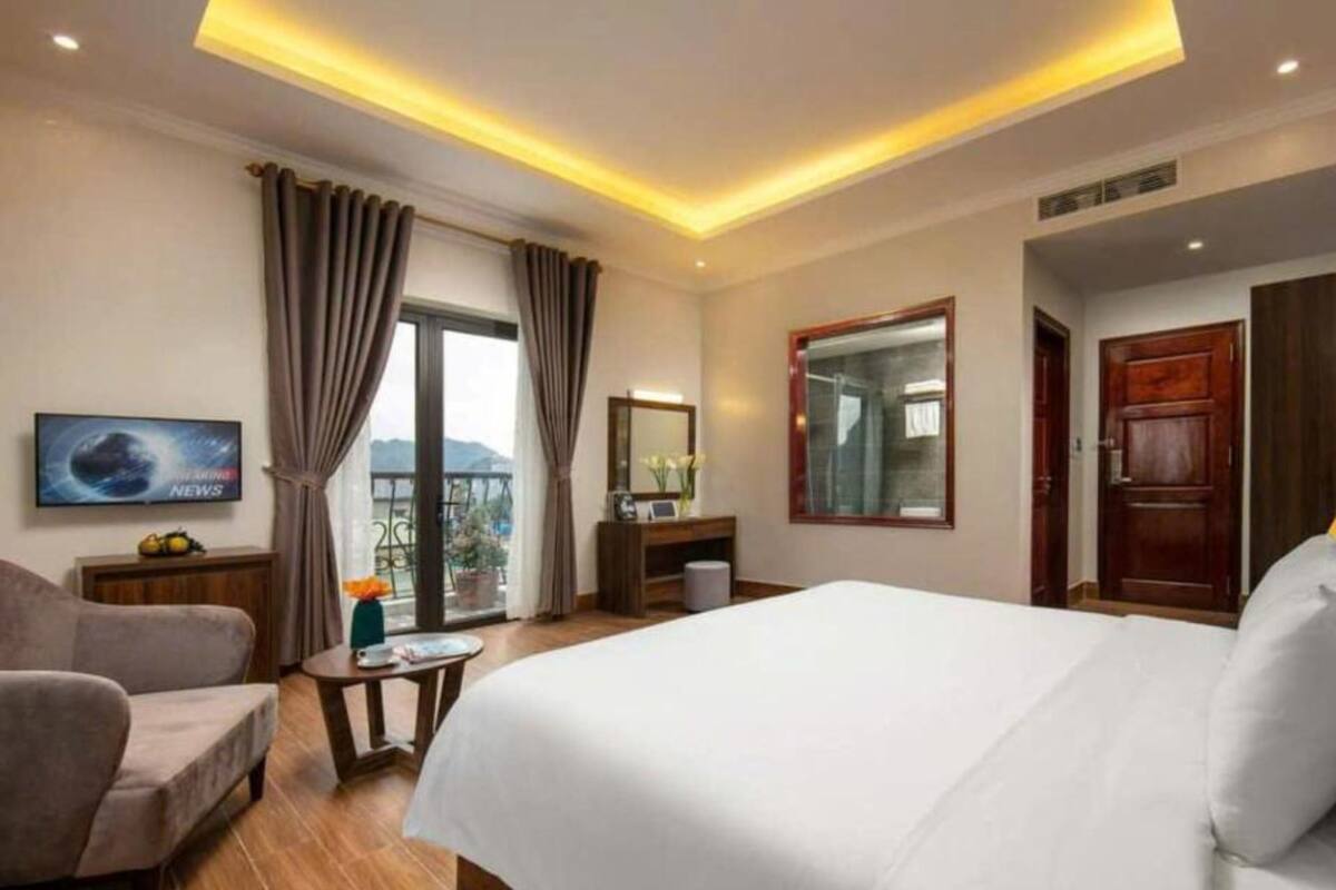 A hotel room with a large bed, a TV on the wall, a desk, and a balcony with curtains.