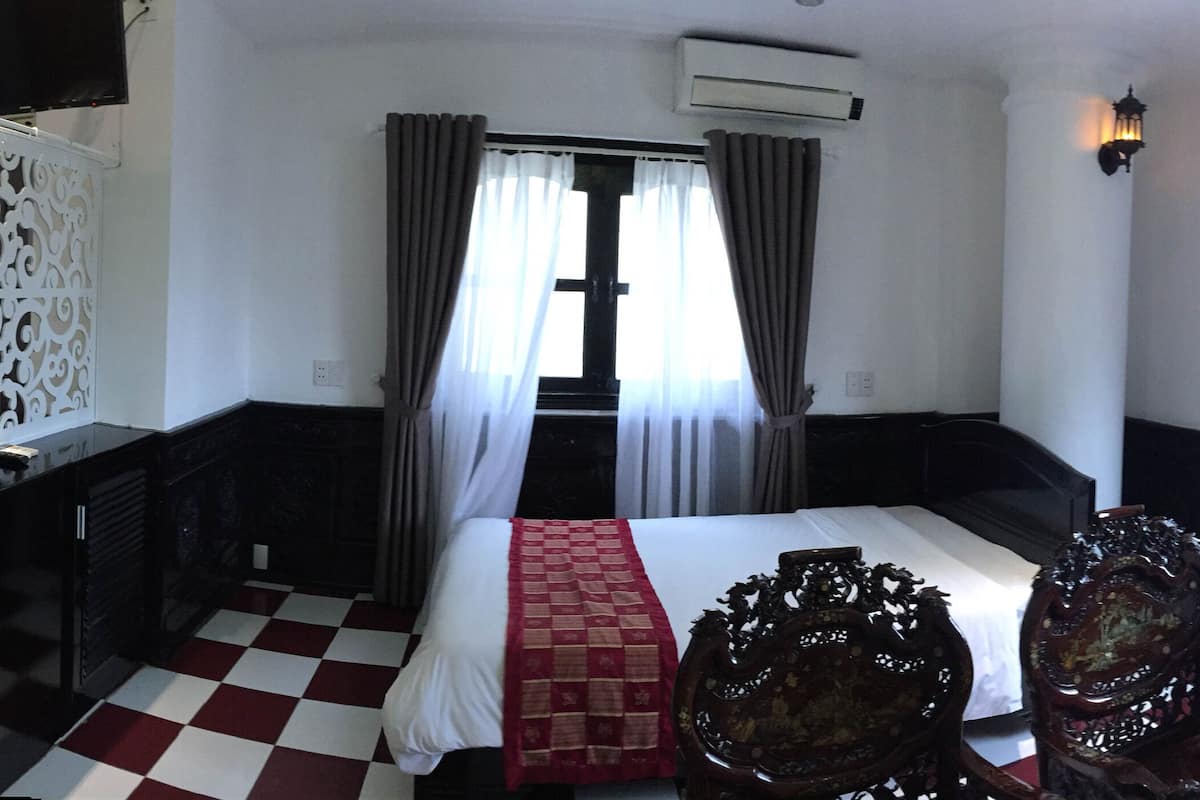 Een hotelkamer met een geruite vloer, een bed met witte lakens, een nachtkastje, een decoratieve kamerdivider, een bank en een traditionele houten stoel.