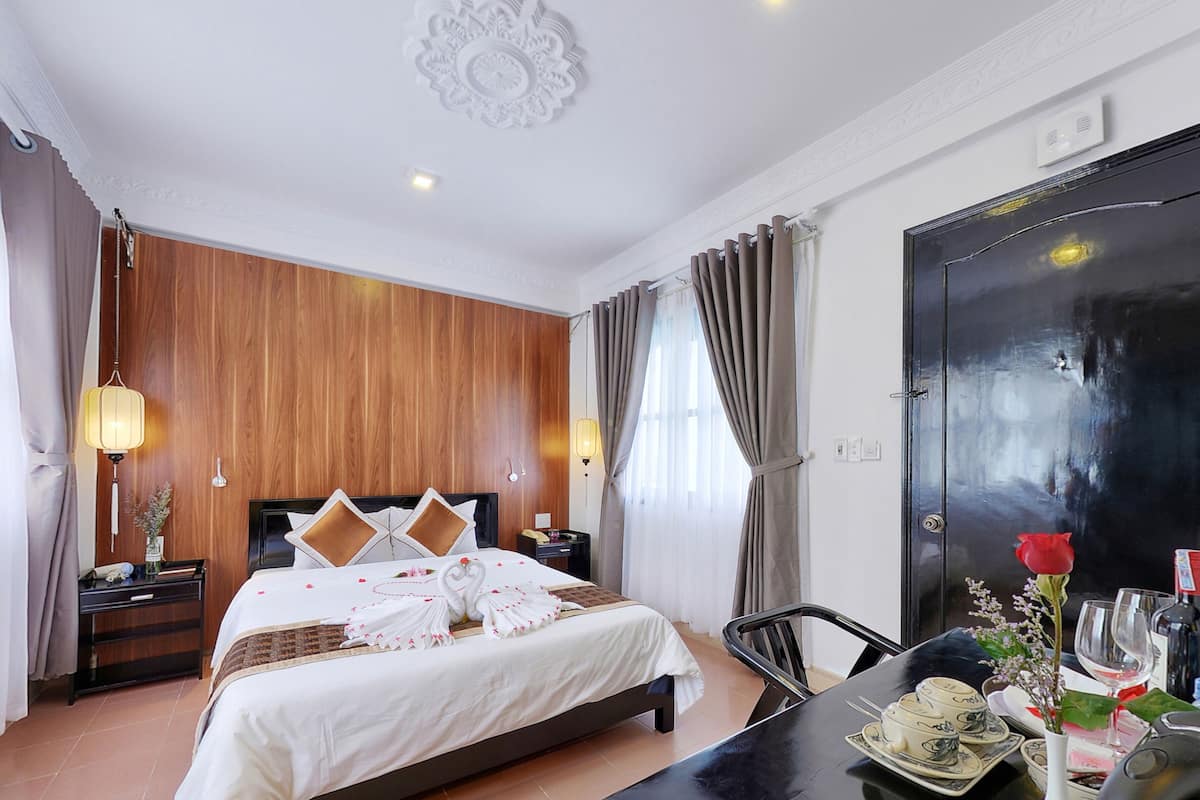 Een hotelkamer met een wand van houten panelen, een bed met wit beddengoed, een zwarte deur, een eettafel met een bloemenvaas en een kroonluchter.