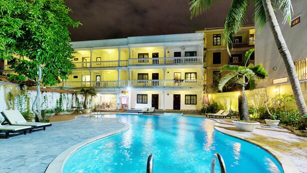 Una piscina iluminada de noche, con sillones de descanso y un edificio de varios pisos al fondo.