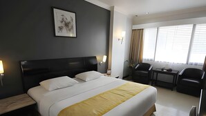 A hotel room with a large bed, a desk, two chairs, a nightstand, and a wall-mounted lamp. - Banjarmasin Internasional (Banjarmasin)
