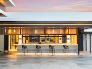 Un espace bar moderne avec un long comptoir et quatre chaises, un comptoir d’accueil bien visible et une vue dégagée sur l’intérieur grâce à la paroi vitrée.