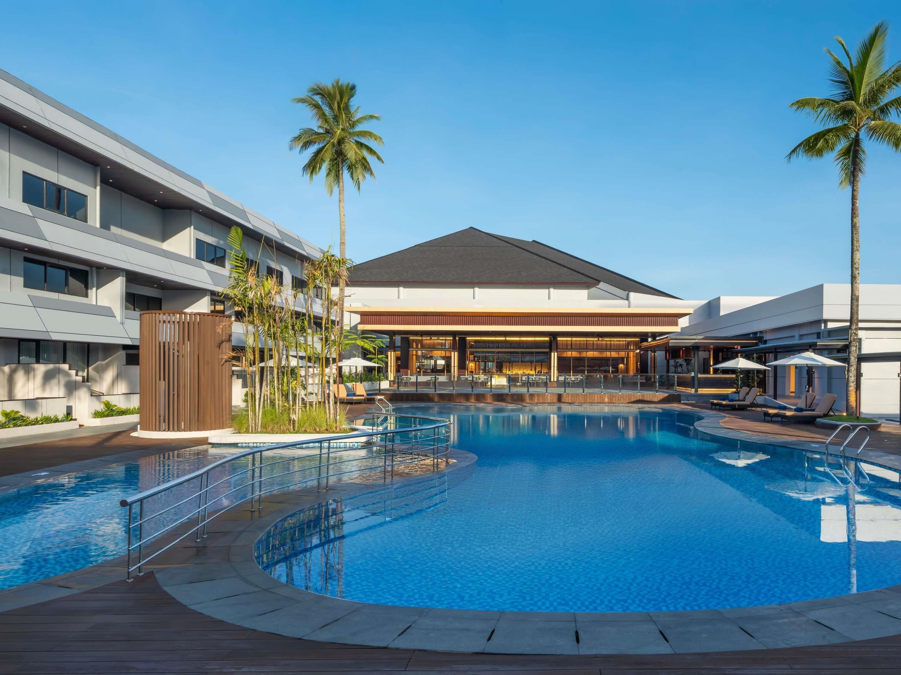 A modern hotel with a central swimming pool, palm trees, and a bar/lounge area.