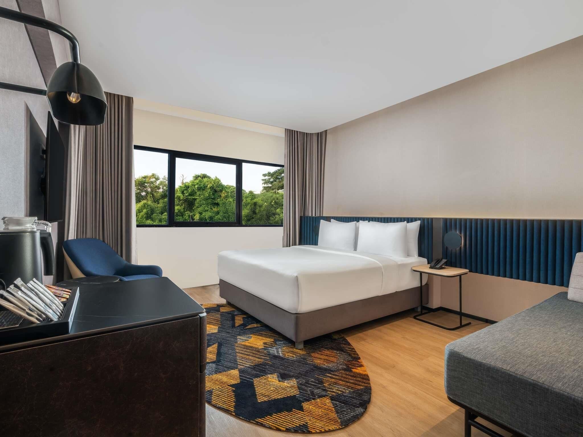 Hotelkamer met een bed, bureau, stoel, bureaulamp en uitzicht op bomen door het raam.