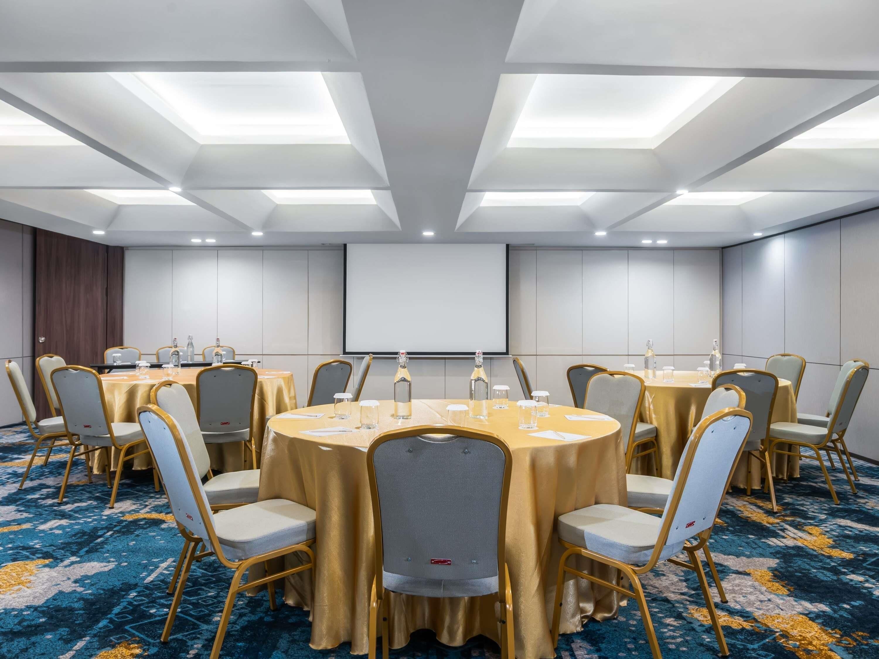 A conference room with round tables, chairs, a projector screen, and a carpeted floor.