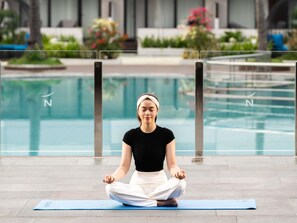 Una persona meditando sobre una esterilla azul junto a una piscina, con vistas despejadas a la piscina y la vegetación circundante.