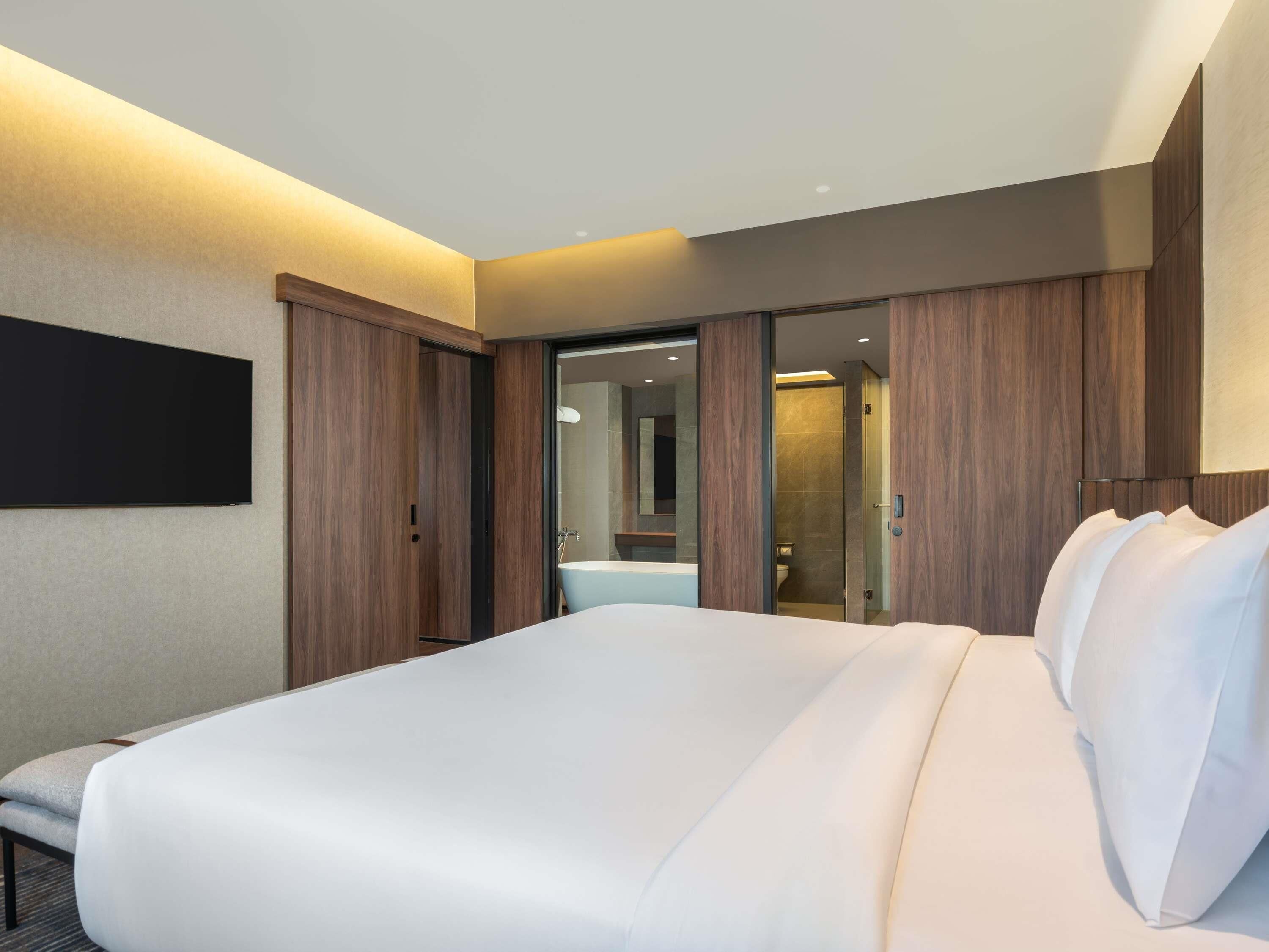 Una habitación de hotel moderna con una cama grande, un armario de madera y un baño visible a través de una puerta abierta.