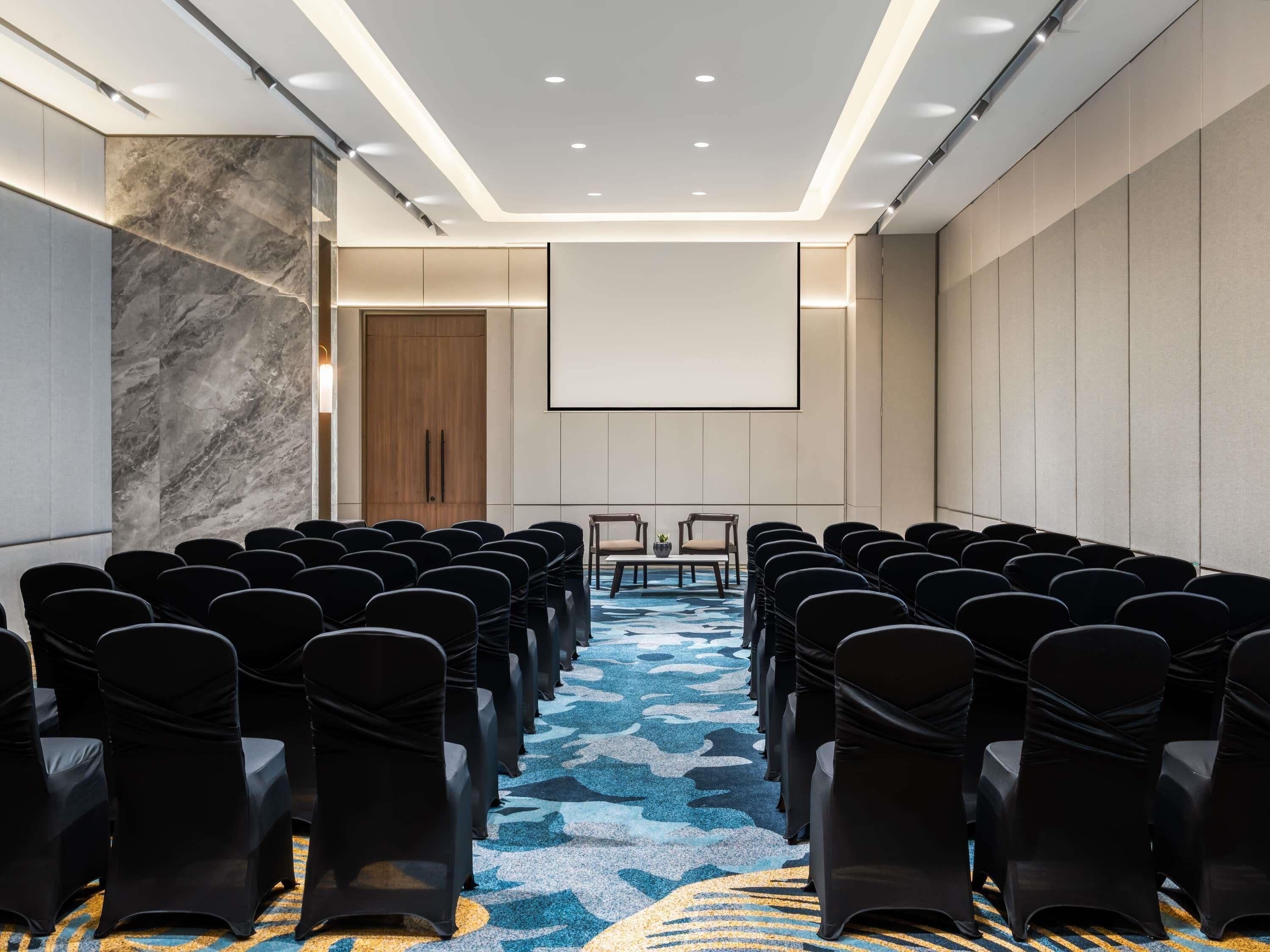 Un salón de conferencias con filas de sillas, una pantalla de proyector y una pared decorativa de mármol.