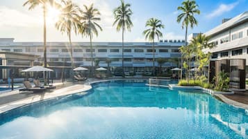 Una piscina con sillas de descanso y sombrillas, rodeada de palmeras y un edificio moderno.