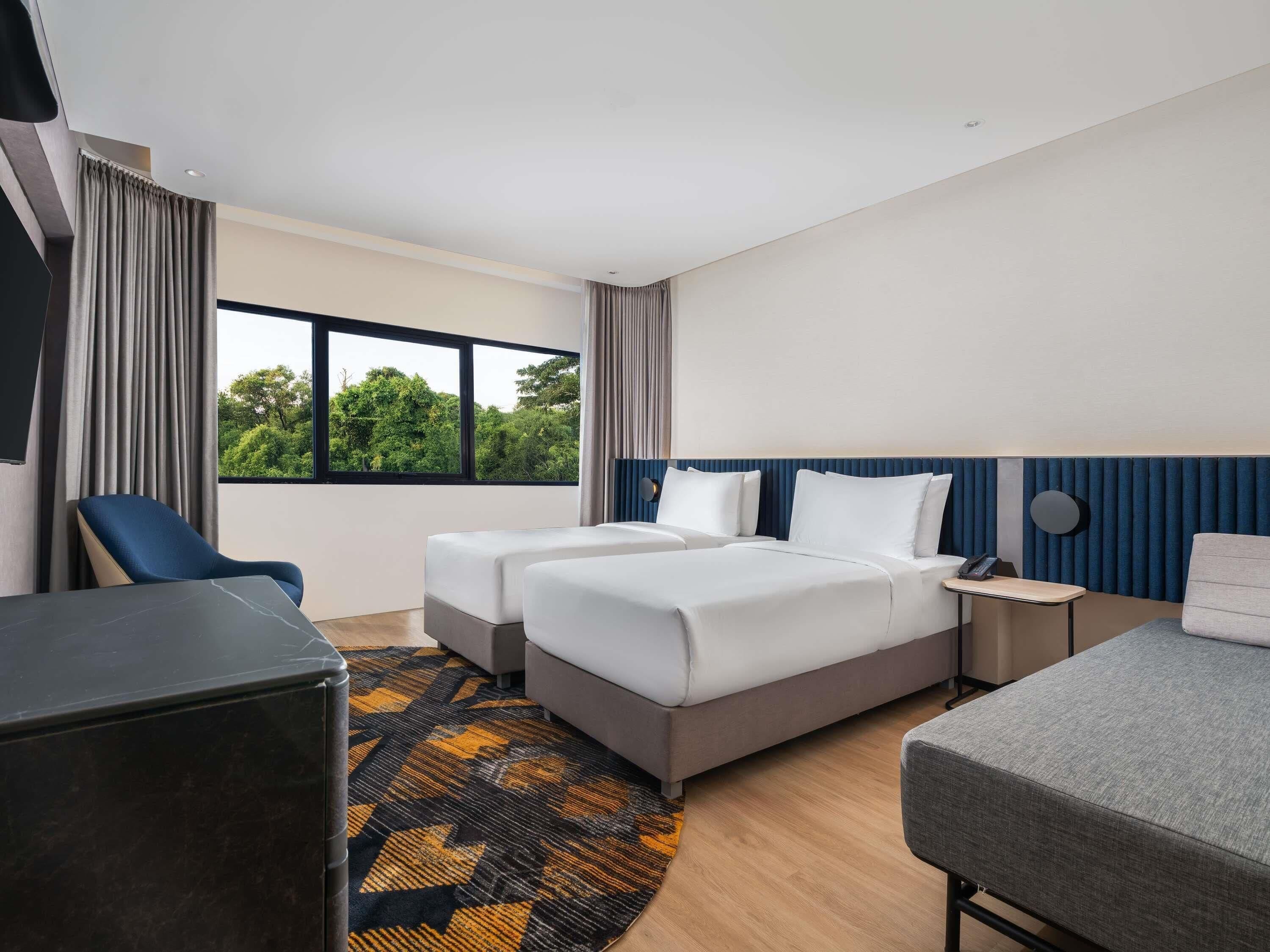 Een hotelkamer met twee bedden, een bureau, een stoel, een televisie en een raam met uitzicht op groen.
