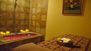 Un cuarto de spa con una bañera llena de pétalos de rosa, una camilla de masaje con un cubrecolchón estampado y un cuadro de flores en la pared.