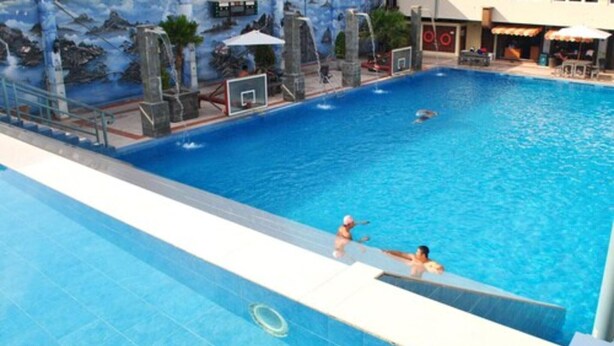 Una piscina con un mural de una cascada y palmeras, un trampolín y un área de descanso con sombrillas.
