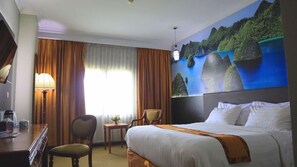 A hotel room with a bed, a desk, a chair, a lamp, and a painting of a coastal landscape. - Grand Victoria Samarinda (Samarinda)