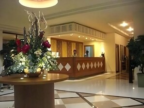 A hotel reception area with a wooden reception desk, a large vase of flowers, and a checkered floor. - Amans (Ambon)