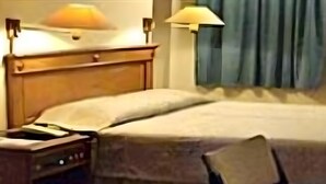 Habitación de hotel con cabecera de madera, mesita de noche con lámpara, una silla y cama con edredón blanco.