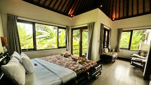 A spacious bedroom with a large bed, a sitting area with a chair and table, and large windows offering a view of lush greenery. - Bali Dream House (Karangasem)