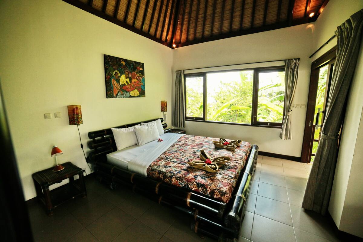 A bedroom with a bed, a painting, a window, and a nightstand.