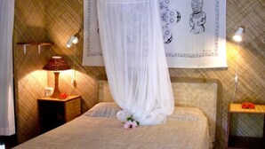 A bed with a canopy, a bedside table with a lamp, and a wall with a tapestry. - Village Temanuata (Bora Bora)