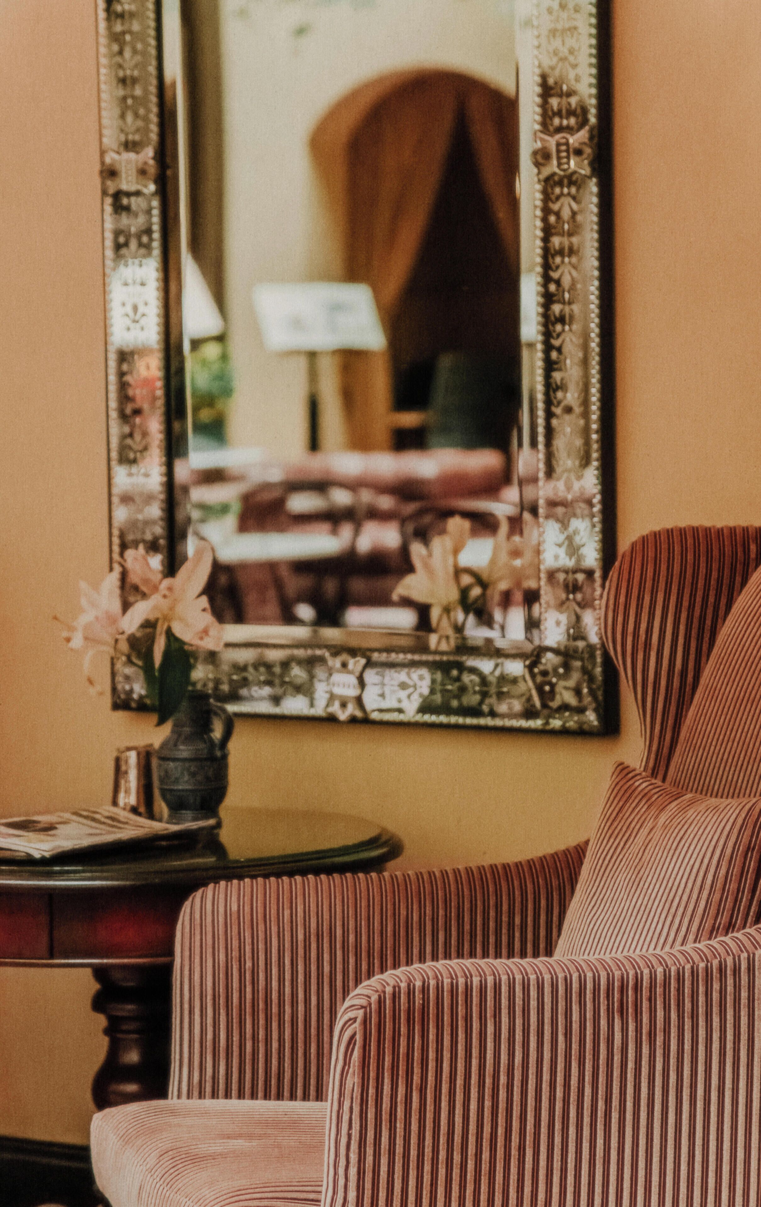 A room with a mirror, a striped armchair, a small table with a vase of flowers, and a lamp.