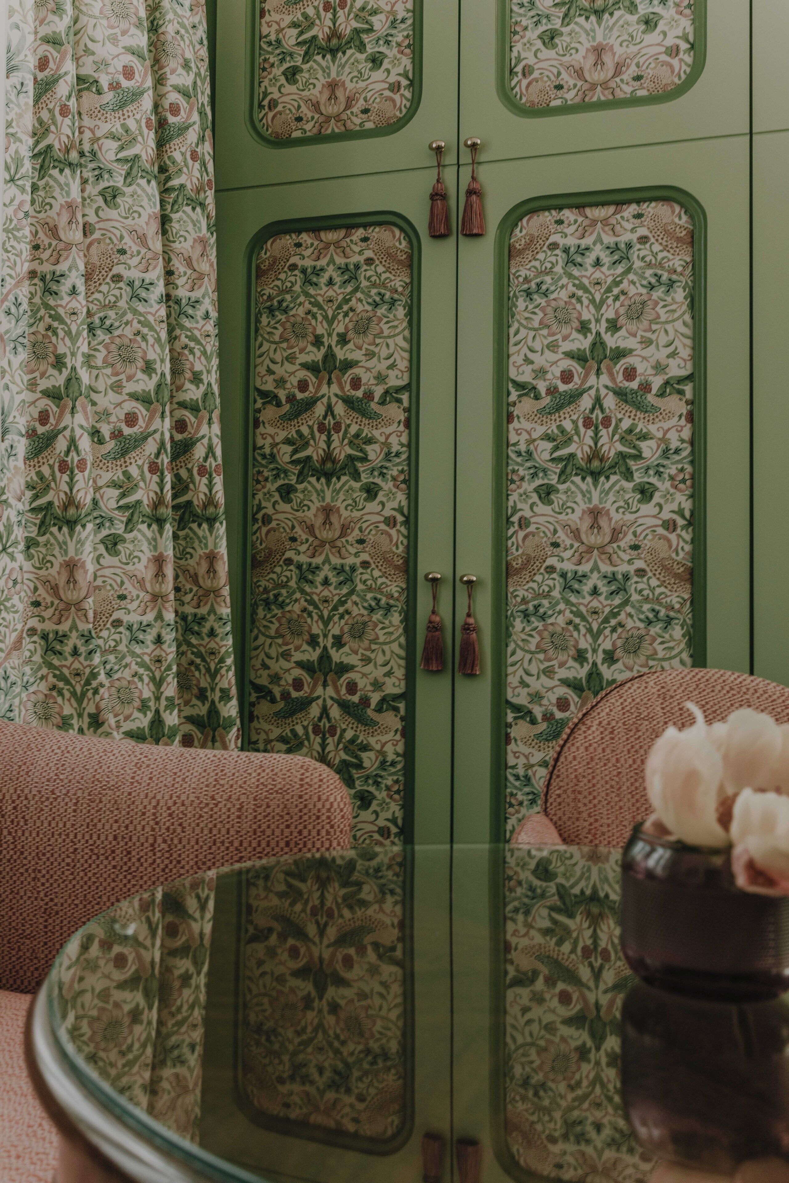 Una habitación con un mueble verde decorado con motivos florales, un espejo redondo y una pequeña mesa con un jarrón.