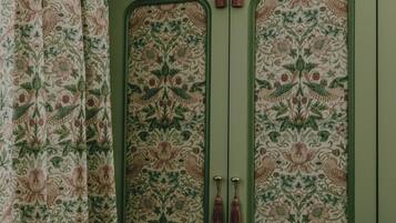 Una habitación con un mueble verde decorado con motivos florales, un espejo redondo y una pequeña mesa con un jarrón.