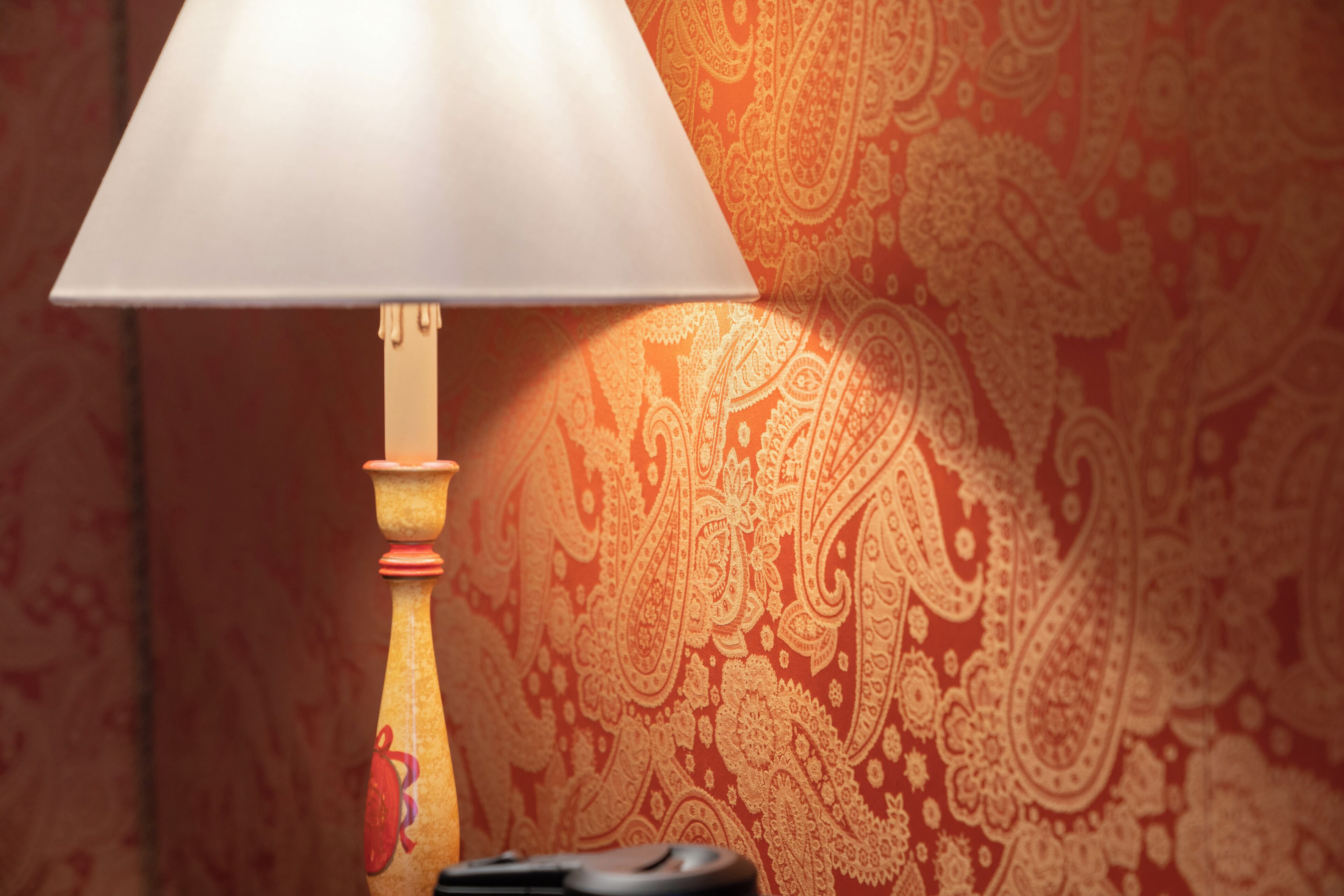 Una lámpara con pantalla blanca y base de madera, colocada sobre una mesa frente a una pared con papel tapiz de estampado floral ornamental.