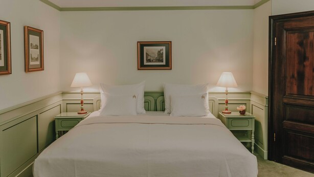 A neatly made bed in a room with two bedside tables, each with a lamp and a framed picture on the wall. - König von Ungarn (Vienna)