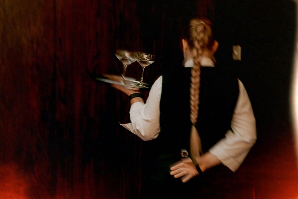 A person holding a tray with a glass and a plate, standing in front of a dark wooden background. - König von Ungarn (Vienna)