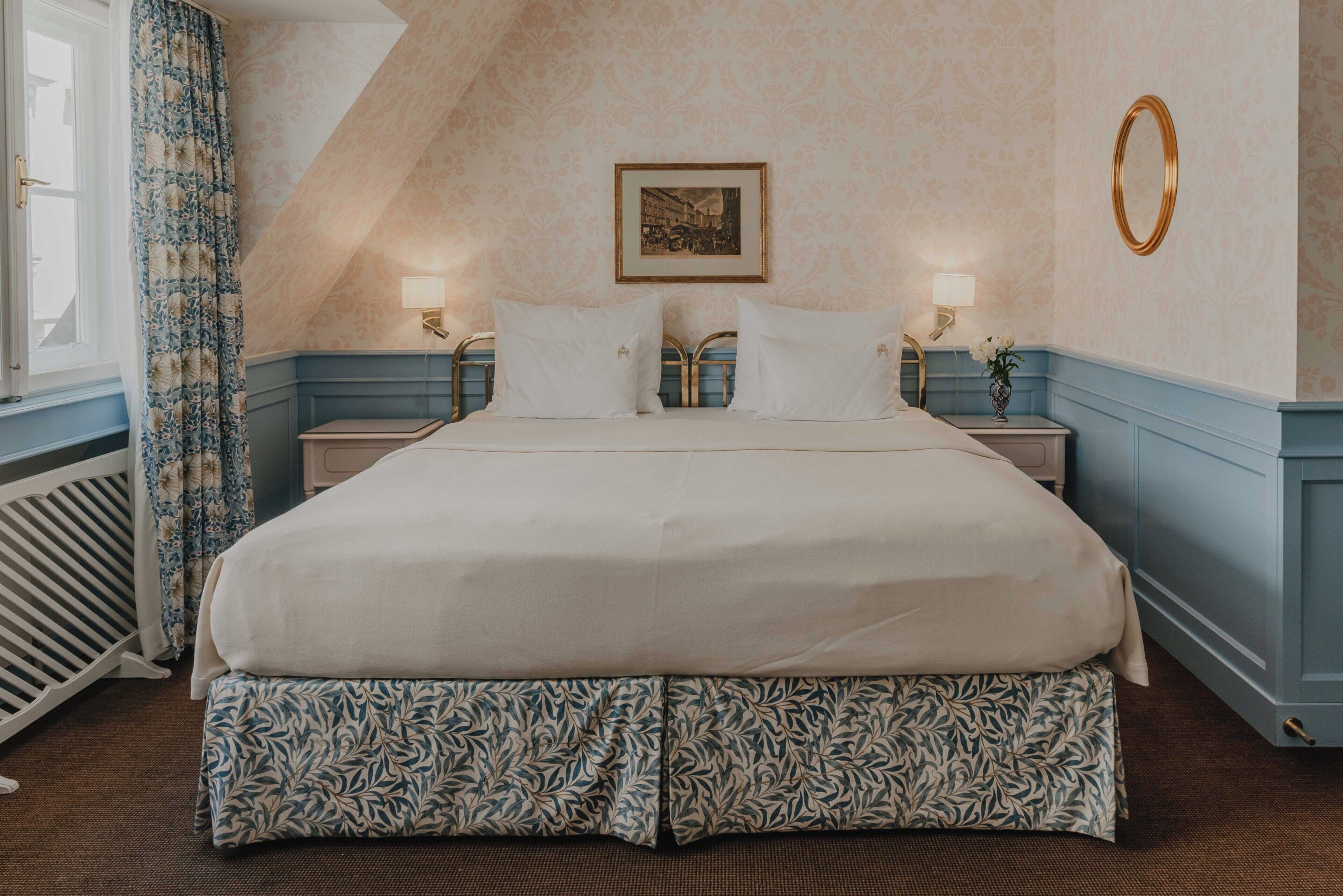 Une chambre d’hôtel avec un grand lit, deux tables de chevet, une lampe fixée au mur, un tableau encadré et un miroir décoratif.