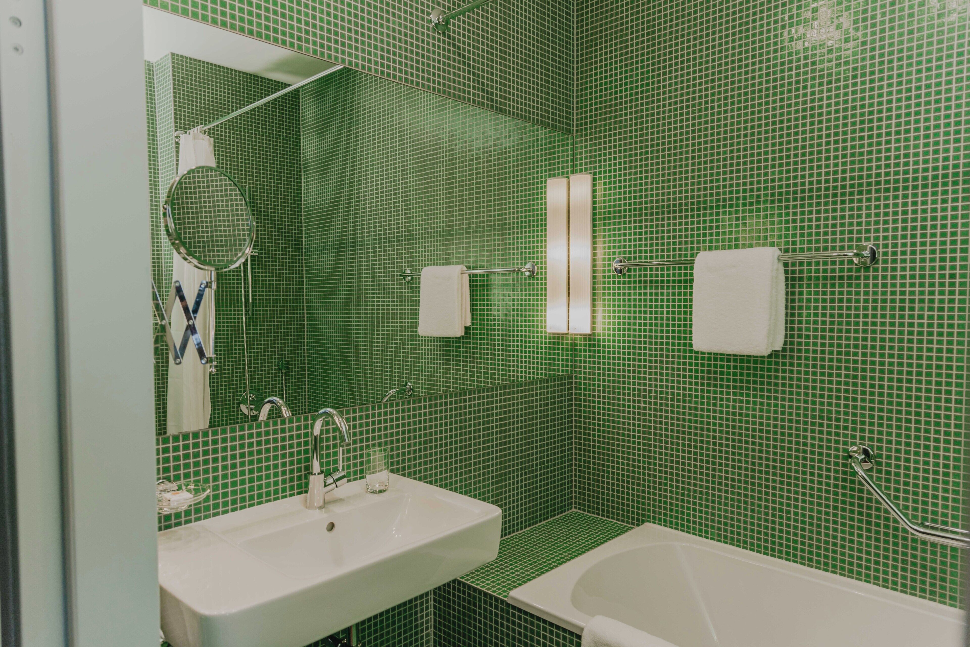 Ein Badezimmer mit grünen Fliesenwänden, einem weißen Waschbecken, einer Badewanne, einem Spiegel und einem Handtuchhalter.