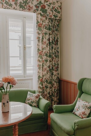 A room with a green armchair, a round table with a vase of flowers, and floral curtains. - König von Ungarn (Vienna)