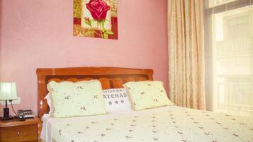 Een hotelkamer met een houten bed, bloemenbedrukking, een nachtkastje met een lamp, een lijst met een roos, en een bordje met de tekst 'HOTEL ATENAS'.
