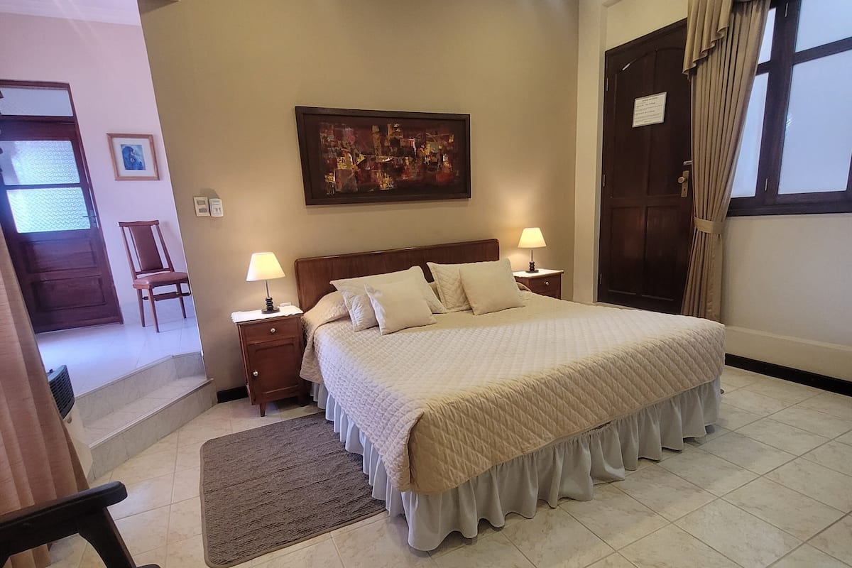 A neatly arranged bedroom with a large bed, two bedside tables with lamps, a framed picture on the wall, and a door with curtains.
