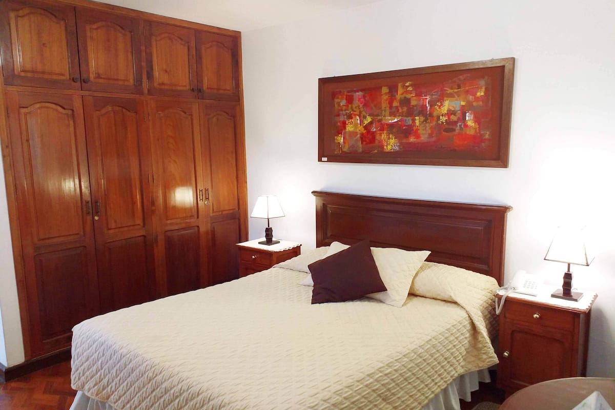 A bedroom with a large wooden wardrobe, a bed with a quilted bedspread, two bedside tables with lamps, and a framed abstract painting on the wall.
