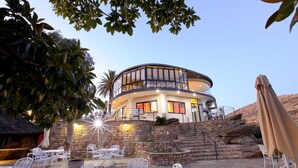 Un edifici modern amb façana corba, zona d'estar a l'aire lliure amb cadires i taules blanques, i una escalinata de pedra que condueix a l'entrada.