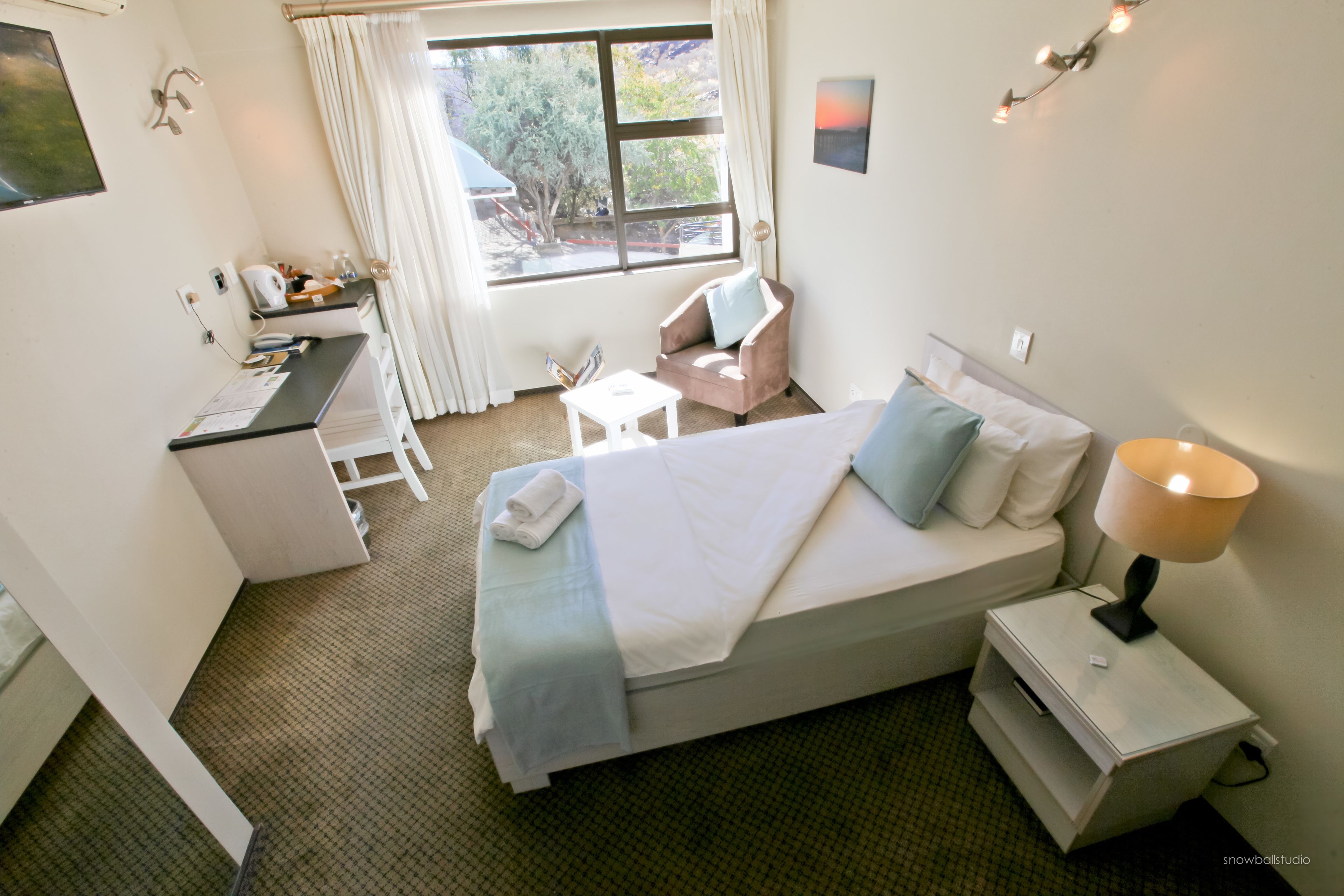 A hotel room with a bed, bedside table, chair, desk, and window.