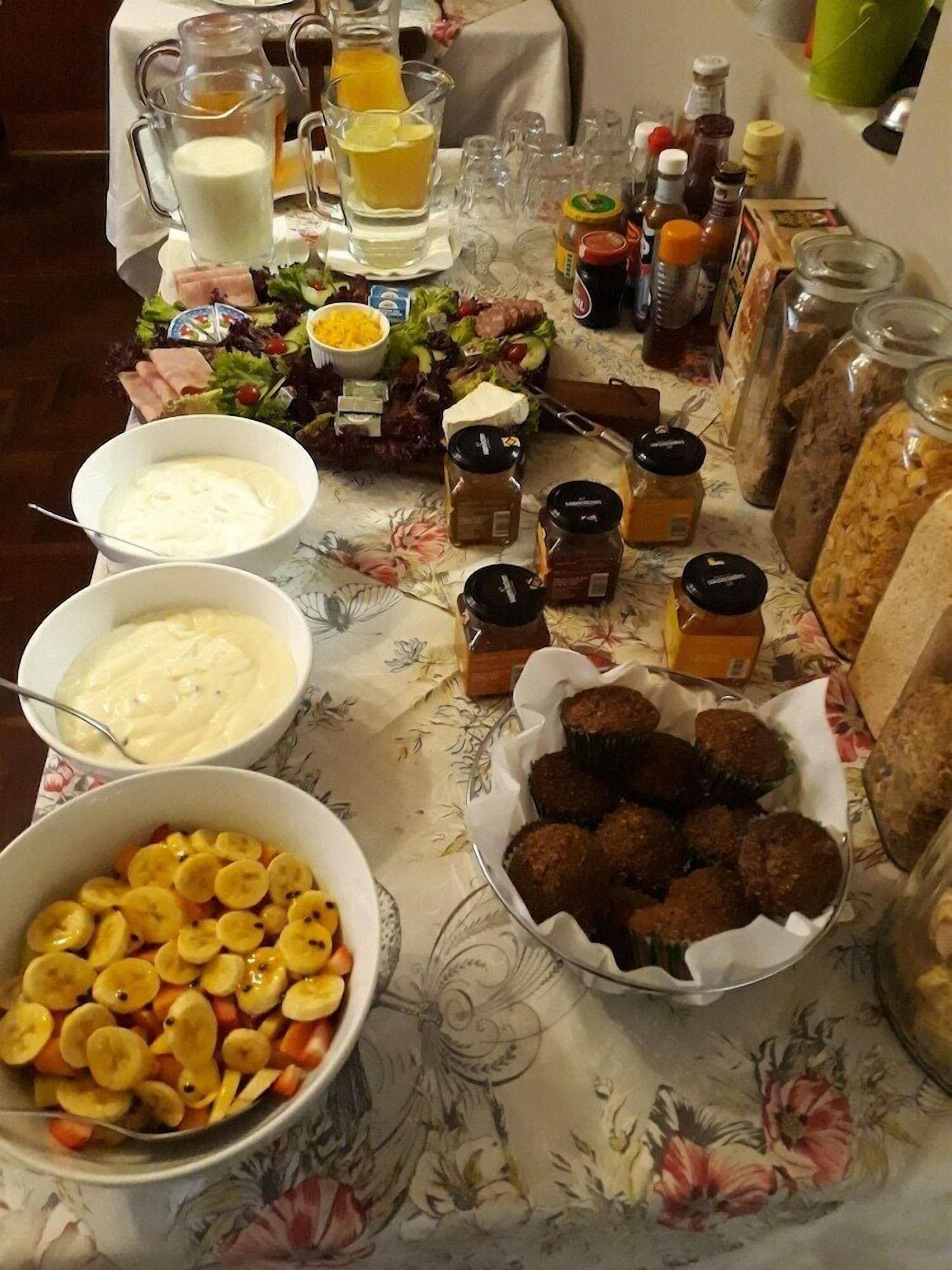 A table with various food items and condiments, including bowls of salad, fruit, and meatballs, as well as bottles of sauces and a pitcher of juice.