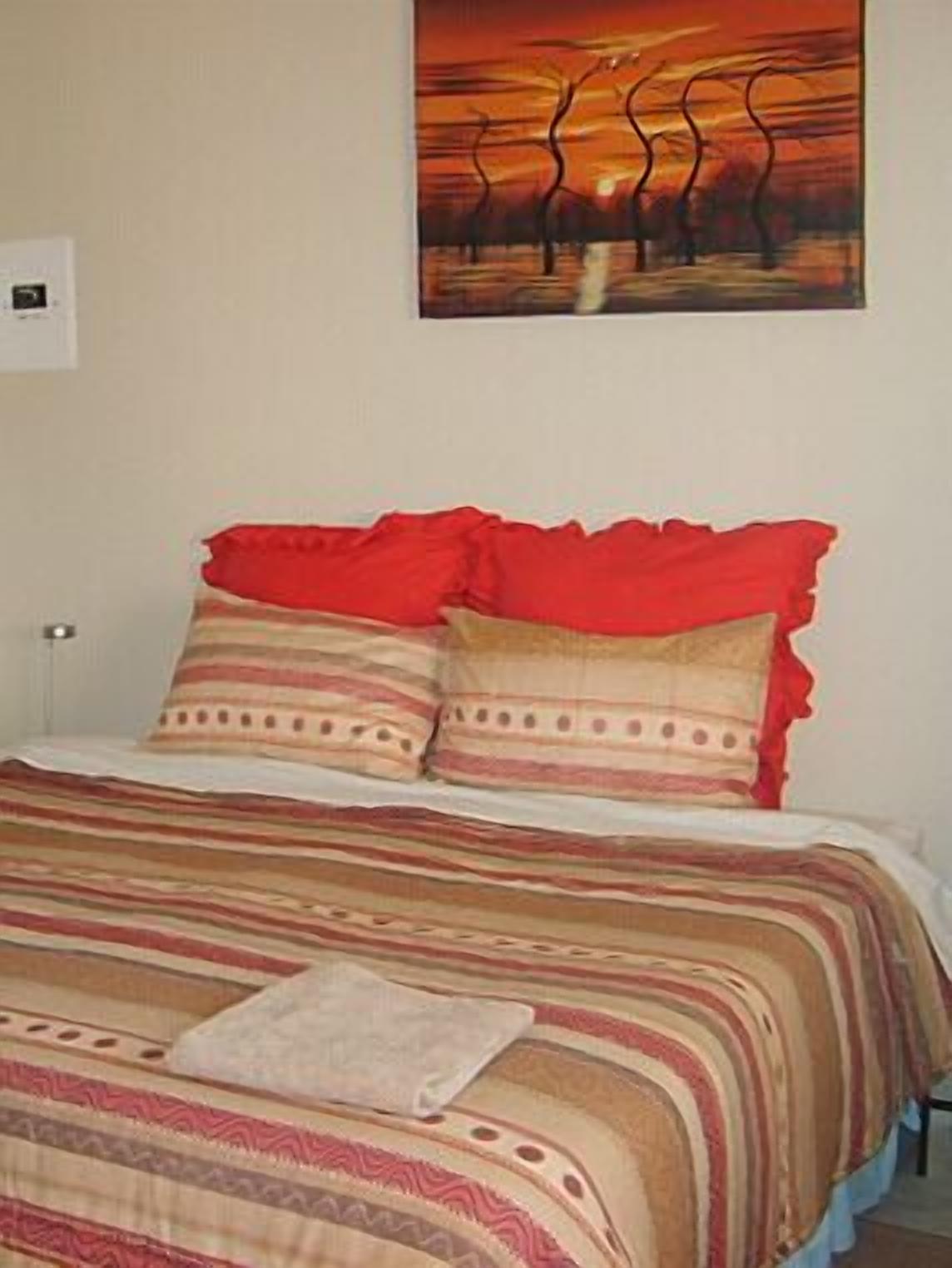 A neatly made bed with striped bedding and red pillows, a painting of a sunset on the wall, and a small bedside table with a lamp.