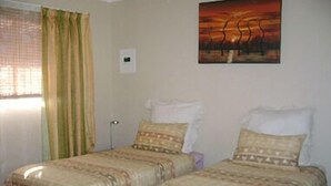 A room with two beds, each with patterned bedding and pillows. There is a window with curtains, a painting on the wall, and a small table with a lamp. - All Are Welcome (Brakpan)